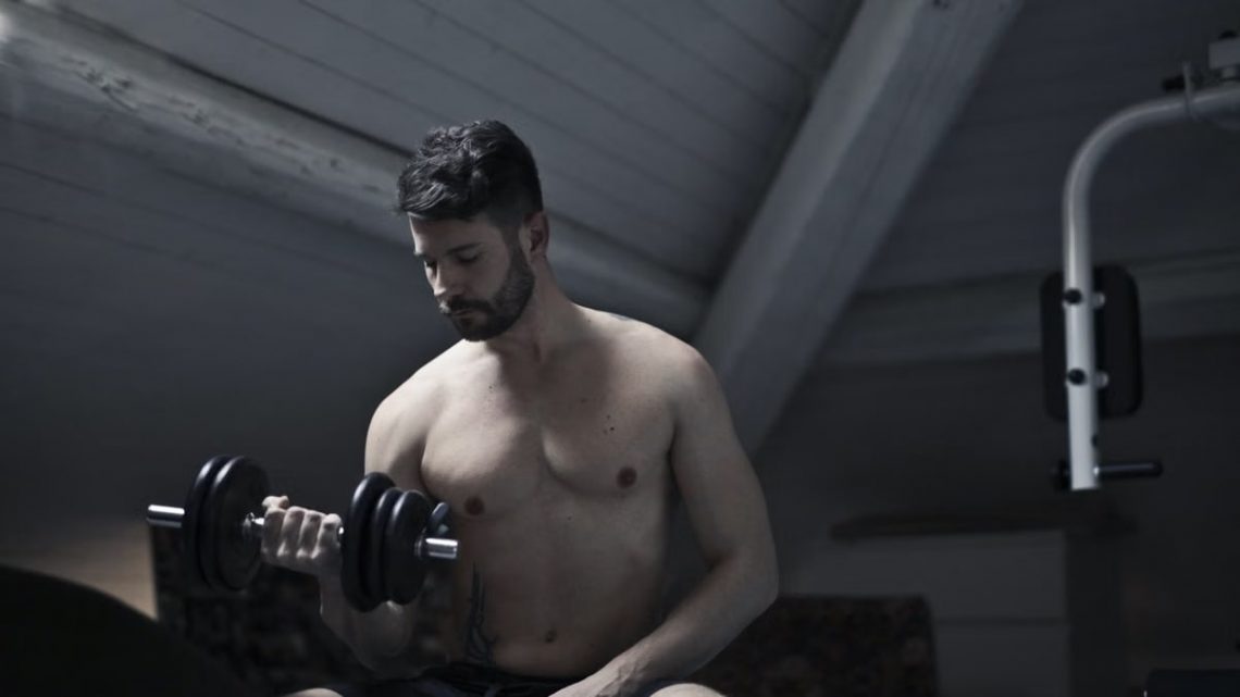 From below of muscular shirtless athletic male sitting on bench and doing bicep curls with dumbbell during intense training in dark studio