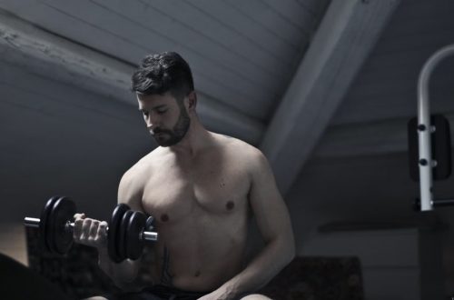 From below of muscular shirtless athletic male sitting on bench and doing bicep curls with dumbbell during intense training in dark studio