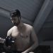 From below of muscular shirtless athletic male sitting on bench and doing bicep curls with dumbbell during intense training in dark studio